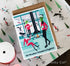 A square greeting card with a kraft envelope, featuring a whimsical winter illustration of three green-skinned characters in a snowy room. The card is placed on top of patterned wrapping paper with a spool of ribbon visible in the lower left.