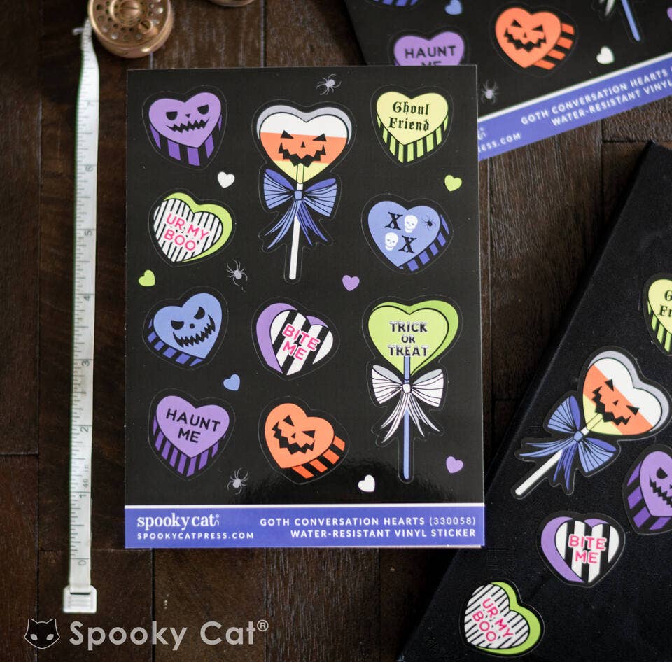 A sheet of Spooky Cat Halloween water-resistant vinyl stickers featuring colorful gothic conversation hearts on a black backing, displayed on a dark wooden table next to a measuring tape.