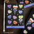 A sheet of Spooky Cat Halloween water-resistant vinyl stickers featuring colorful gothic conversation hearts on a black backing, displayed on a dark wooden table next to a measuring tape.