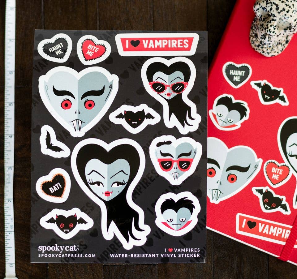 A sheet of vampire-themed water-resistant vinyl stickers on a dark wood background, next to a red sticker album cover. The stickers feature cartoon vampire faces, bats, and heart-shaped sweets with text like &
