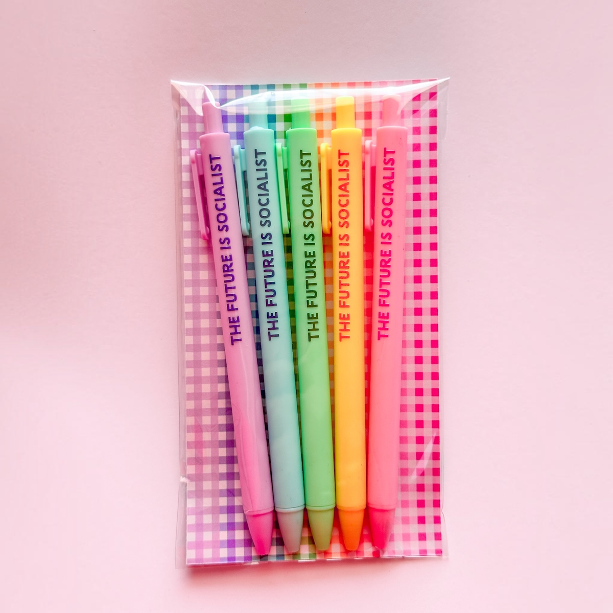 A set of six pastel-colored pens in a clear plastic sleeve with a pink and white gingham-patterned backing card. Each pen is printed with the text &