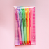 A set of six pastel-colored pens in a clear plastic sleeve with a pink and white gingham-patterned backing card. Each pen is printed with the text &