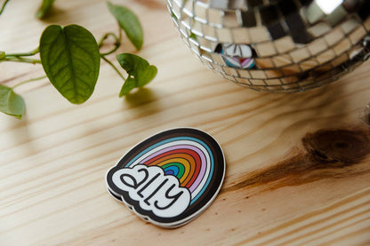 Sticker with rainbow design and text on a wooden surface with a disco ball and plant.