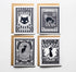 Four Halloween-themed postage stamp greeting cards arranged in a 2x2 grid on a white background, each with a kraft envelope behind it. The stamps feature black cats, vintage typography, and designs in black, white, and navy tones.