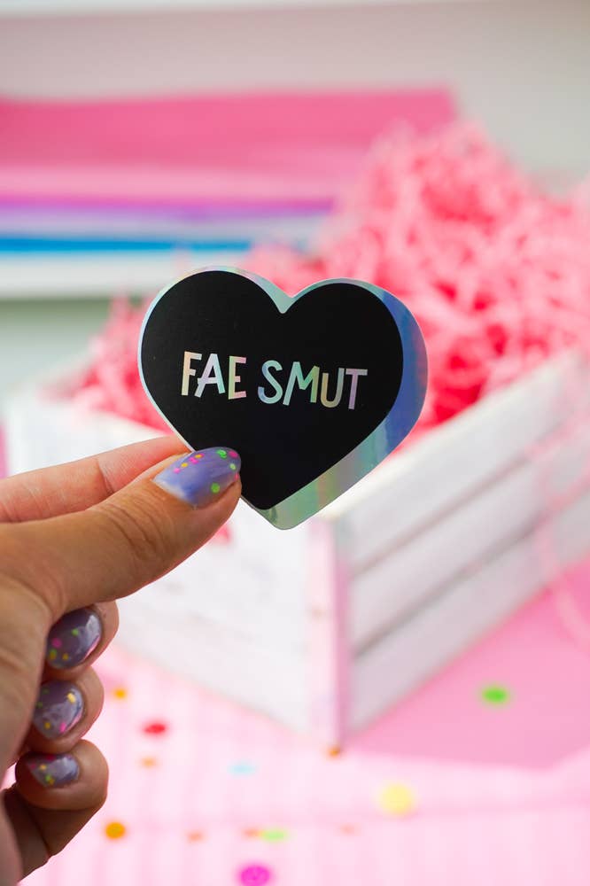 A hand with glittery purple nail polish holds a holographic heart-shaped sticker that reads &