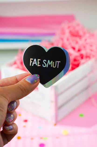 A hand with glittery purple nail polish holds a holographic heart-shaped sticker that reads &