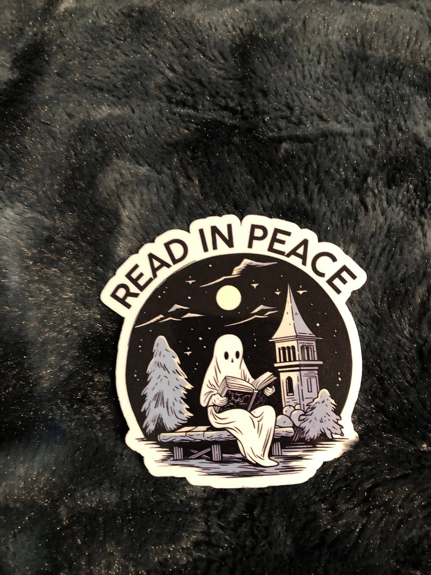 A die-cut sticker featuring a ghost reading a book under a full moon near a church tower, with the words &