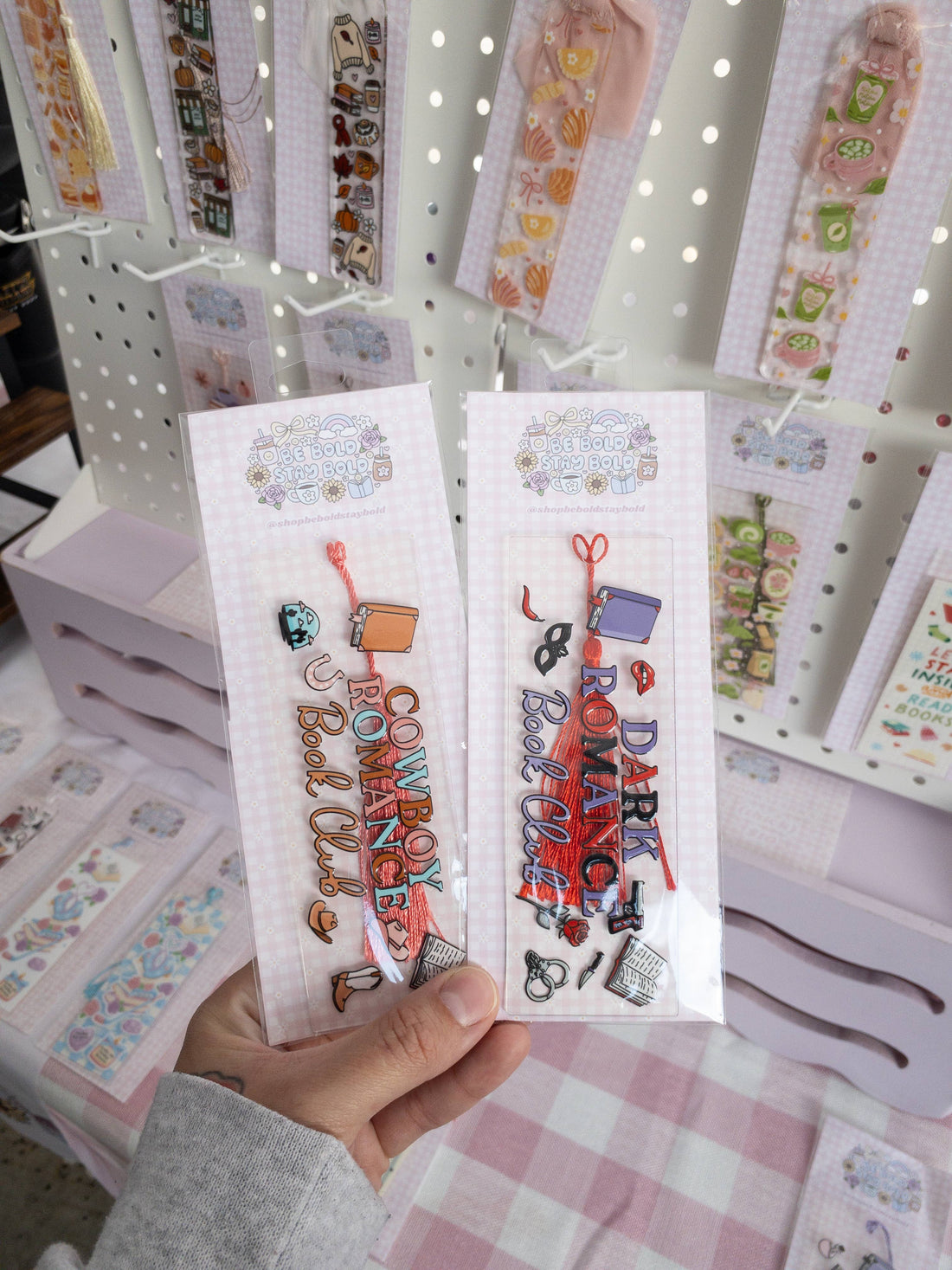 Two bookmarks with colorful text and designs held by a hand, with a display of more bookmarks in the background.