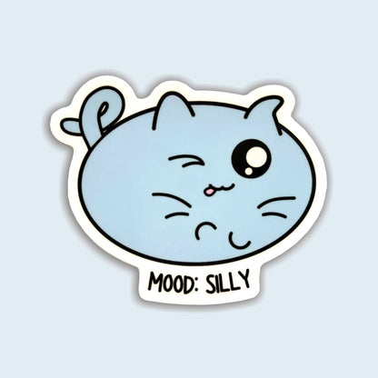 A die-cut sticker featuring a light blue cartoon cat face with one winking eye and the text &