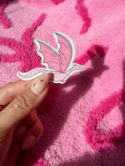 A hand with decorated fingernails holds a die-cut sticker of a cute pink dragon against a textured hot pink blanket.