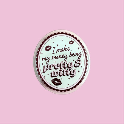 A circular button with the text "I make my money being pretty &amp; witty" printed on it, set against a pink background.