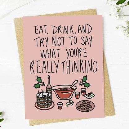 A pink greeting card with a humorous quote and a holiday-themed illustration, displayed on top of a brown kraft envelope against a white wooden background.