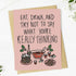 A pink greeting card with a humorous quote and a holiday-themed illustration, displayed on top of a brown kraft envelope against a white wooden background.