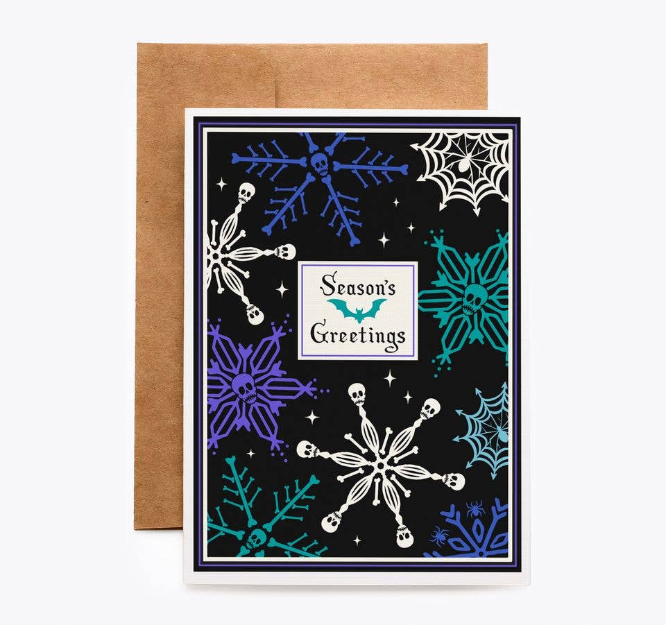 A Halloween-themed greeting card with a brown kraft envelope behind it. The card has a black background decorated with white, blue, and teal snowflakes, spiderwebs, stars, and a central white label that reads &