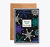 A Halloween-themed greeting card with a brown kraft envelope behind it. The card has a black background decorated with white, blue, and teal snowflakes, spiderwebs, stars, and a central white label that reads &