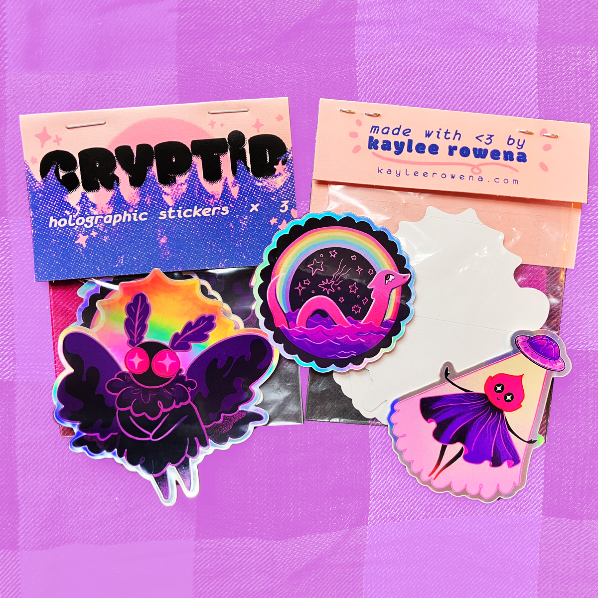 A set of colorful holographic stickers displayed on a purple plaid background. The stickers include a purple winged creature with pink eyes, a rainbow with a pink snake, and a slice of cake with a face, alongside branded backing cards reading &