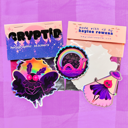 A set of colorful holographic stickers displayed on a purple plaid background. The stickers include a purple winged creature with pink eyes, a rainbow with a pink snake, and a slice of cake with a face, alongside branded backing cards reading &