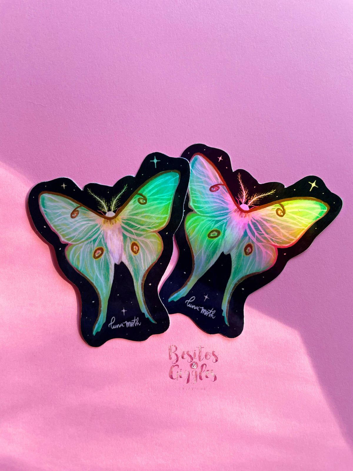Two butterfly-shaped stickers with iridescent pastel wings on a black starry background, displayed on a pink surface with the text &