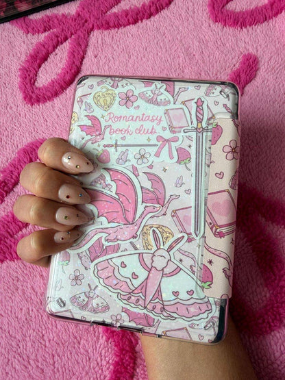 A hand with decorated nails holds a slim hardcover notebook or journal featuring a pink and white anime-style illustration with the text &