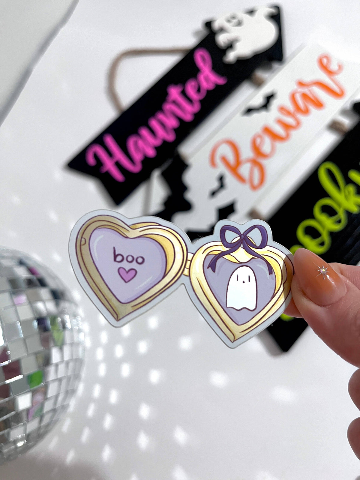 A hand holds a pair of novelty heart-shaped sunglasses with a gold frame and purple lenses, one lens showing the word &