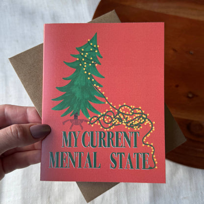 A hand holds a coral-pink greeting card featuring a green Christmas tree with tangled yellow string lights and the text &