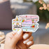 Hand holding a colorful sticker with LGBTQIA+ symbols and text.