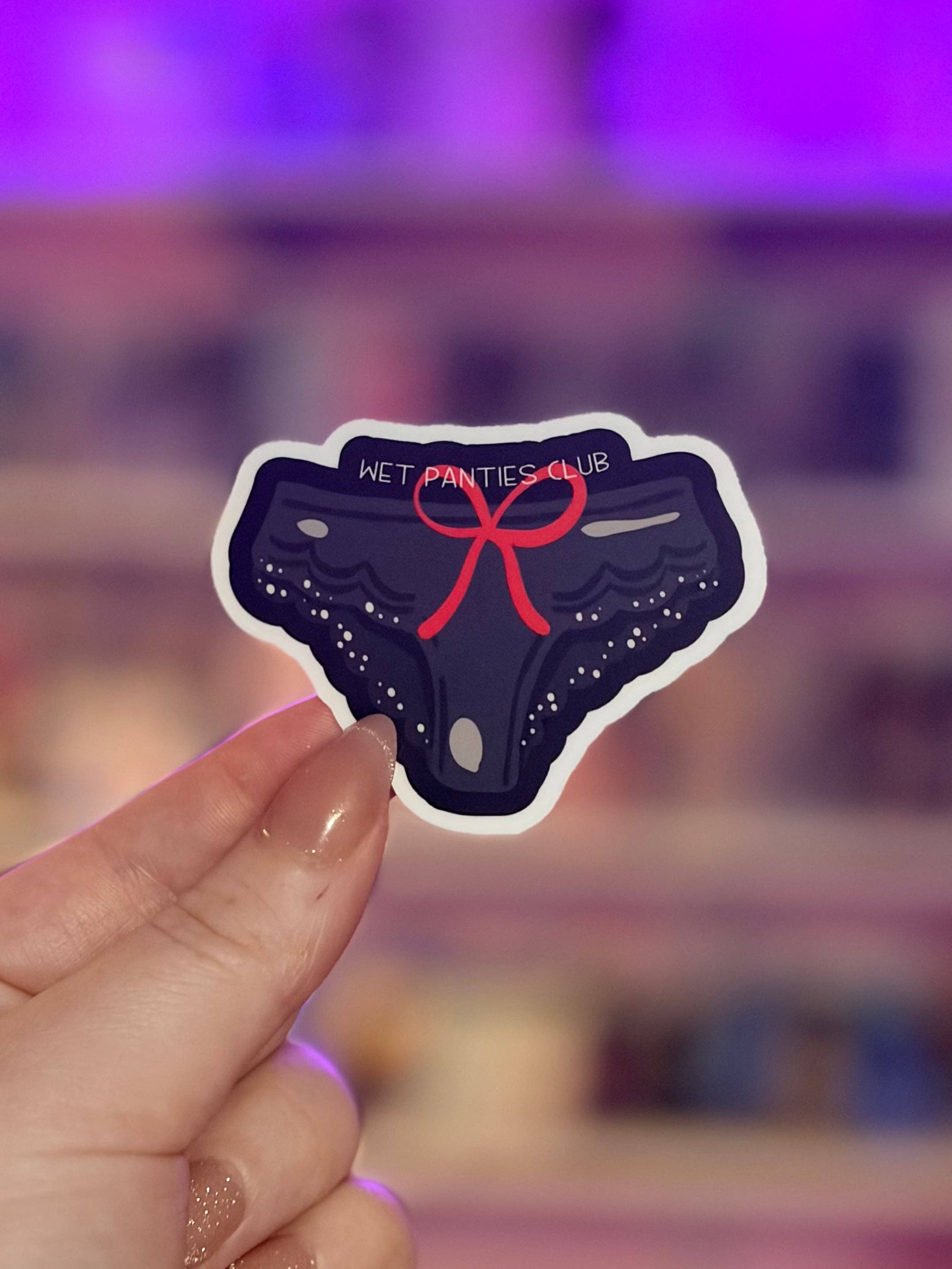 Sticker of a pair of panties with a red bow held by a hand against a blurred background.
