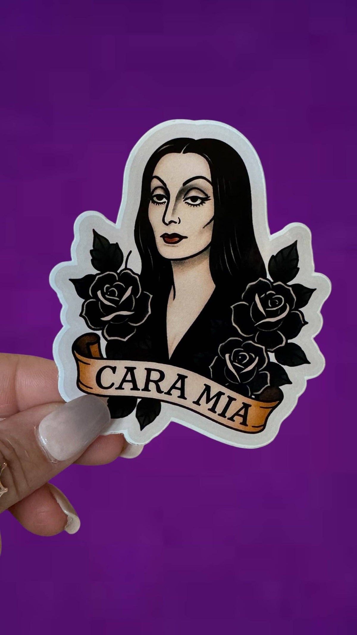 A hand holds a die-cut sticker featuring an illustrated woman with long black hair, black roses, and a banner reading &