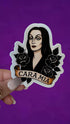 A hand holds a die-cut sticker featuring an illustrated woman with long black hair, black roses, and a banner reading &