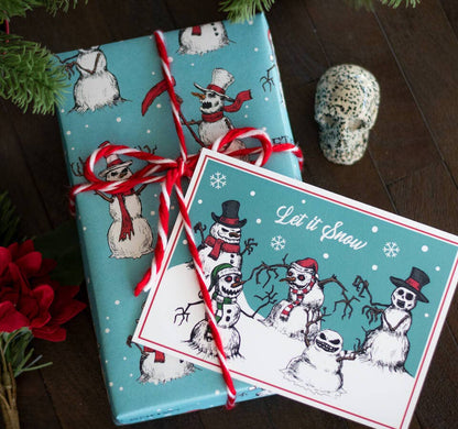 A wrapped gift box and matching greeting card featuring a winter snowman theme, tied with a red-and-white candy cane ribbon on a dark wooden surface with decorative greenery and a skull prop.