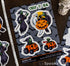 A sheet of Halloween vinyl stickers featuring grim reaper, witch, ghost, and pumpkin car designs on a dark gray background with bats. The sticker sheet is photographed on a dark wood surface with a ruler and skull props nearby.