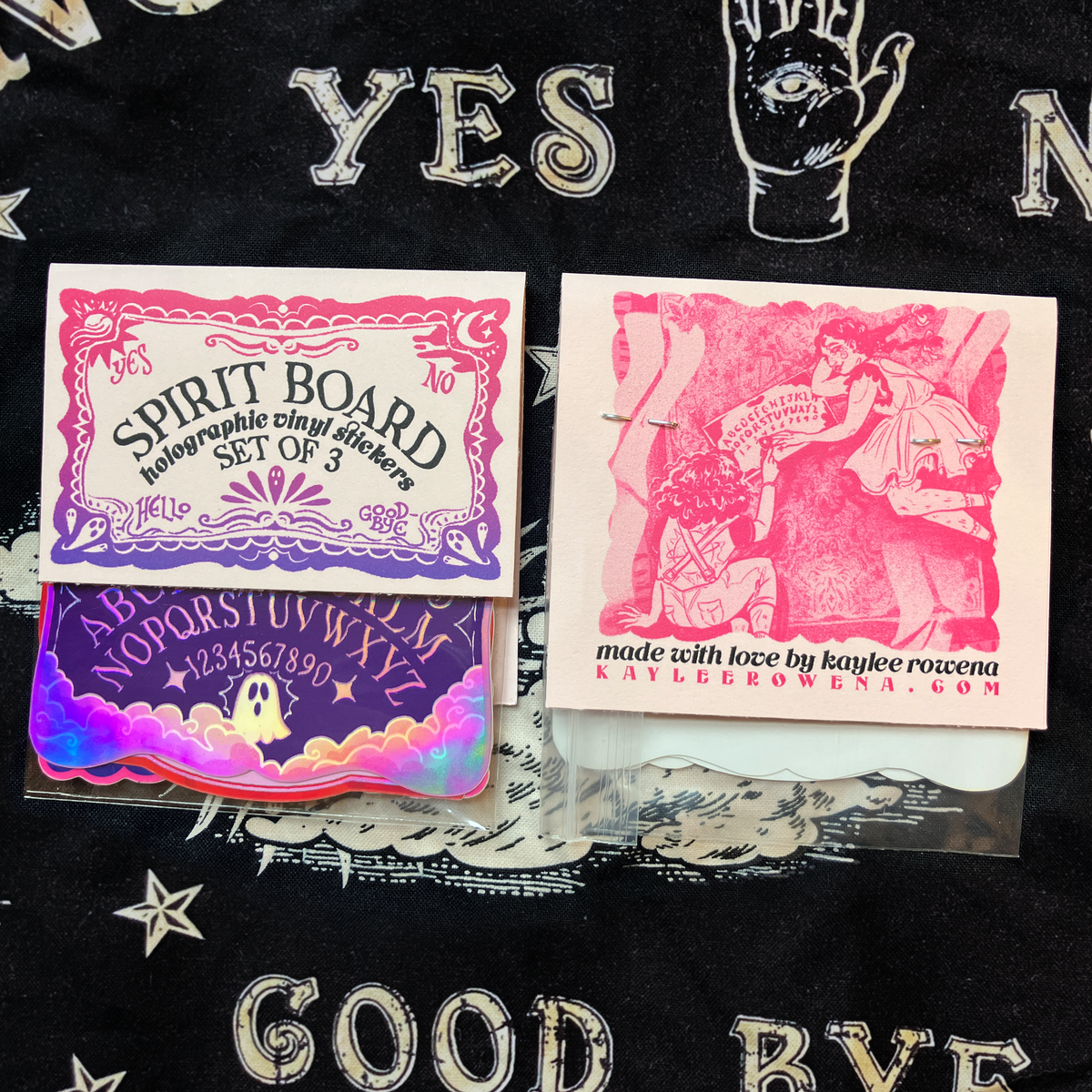 A set of three occult-themed holographic vinyl stickers displayed on a black fabric background. The stickers feature gothic designs including a &