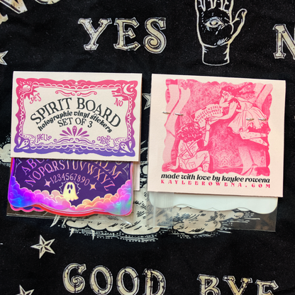 A set of three occult-themed holographic vinyl stickers displayed on a black fabric background. The stickers feature gothic designs including a &