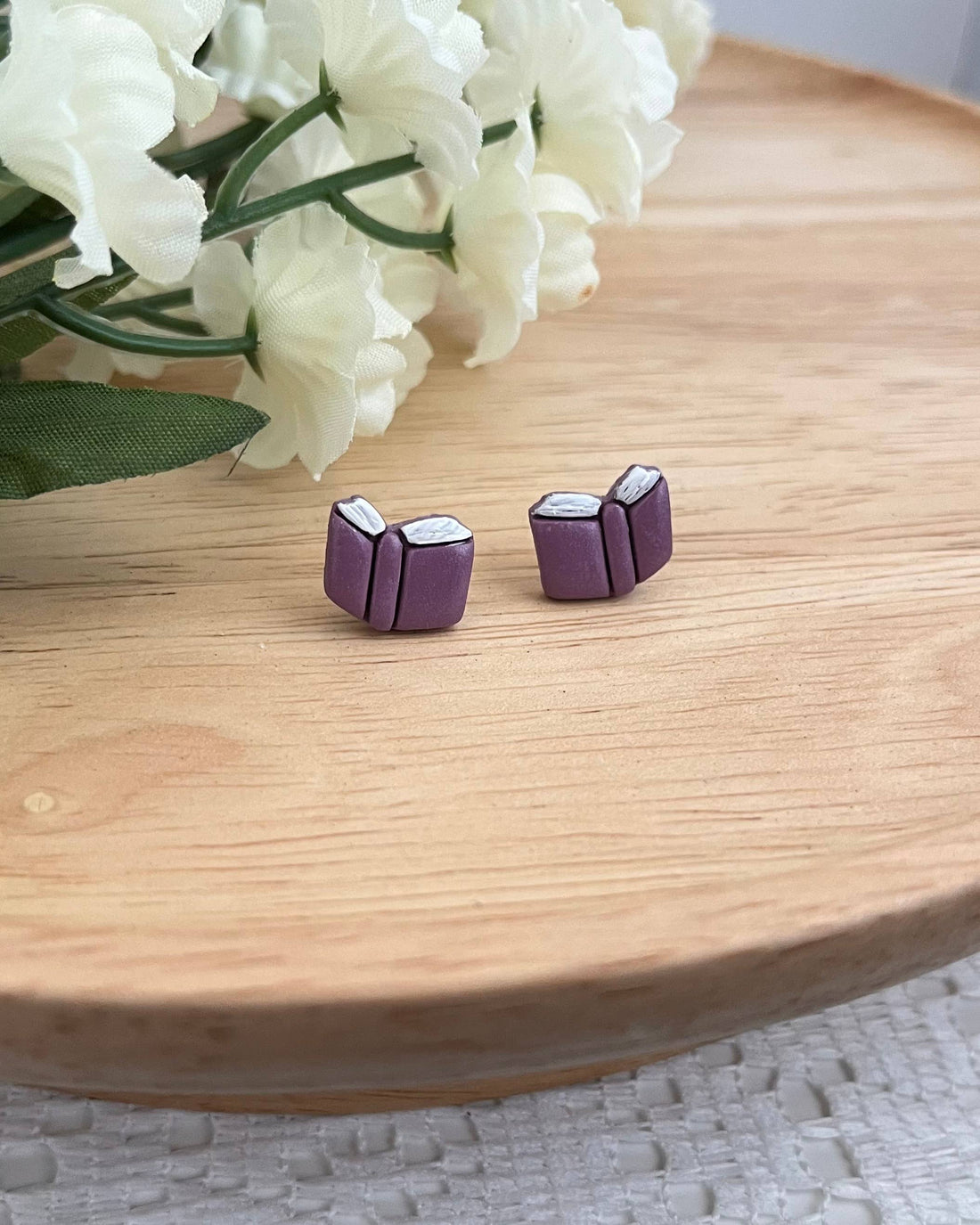 Purple book-shaped earrings on a wooden surface with white flowers in the background