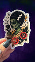 A hand with dark green nail polish holds a die-cut sticker featuring a traditional tattoo-style illustration of a green-skinned woman with black hair, red roses, and a lightning bolt, set against a dark purple textured background.