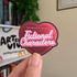 Heart-shaped sticker with text held by a hand in front of a bookshelf.