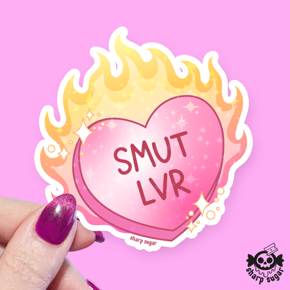 A die-cut sticker held by a hand with glittery purple nail polish. The sticker features a pink heart with the text &