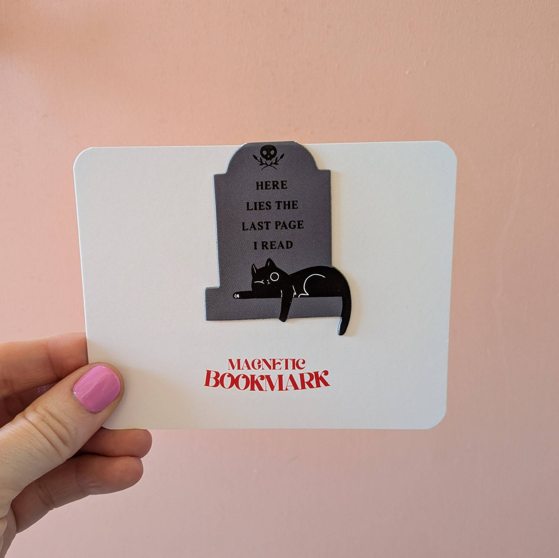 Magnetic bookmark with cat design and text held by a hand on a pink background