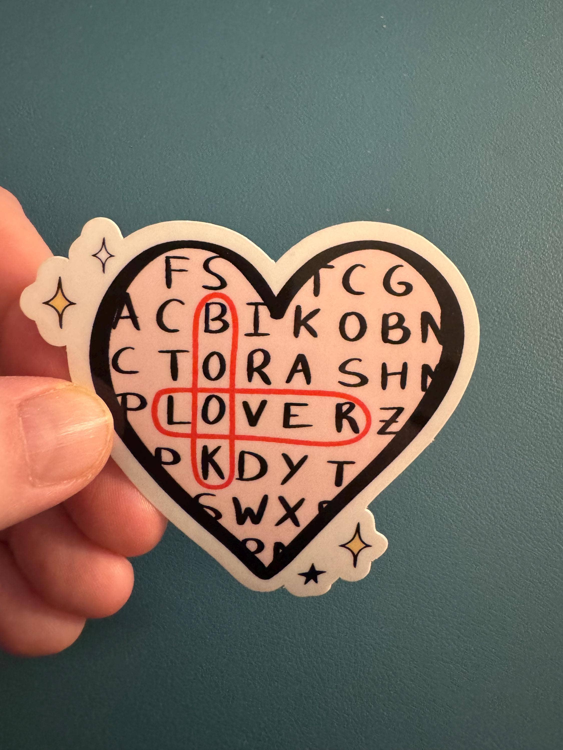 A hand holding a heart-shaped sticker with a crossword puzzle design. The sticker features a black heart outline, a light pink background, black letters spelling out &