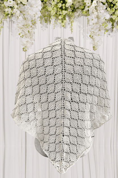 An organic merino/cotton beaded shawl is hand knit in a stunning leaf lace pattern, with subtle coordinating Czech glass beads. It is shown over a wedding dress, with a white fabric draping in the background with white flowers draped over it.