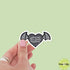 Heart-shaped sticker with bat wings and text held by a hand against a light green background