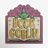 A square decorative graphic or sticker featuring the words “BOOK GOBLIN” in large yellow letters over an open book, with a green goblin face and blue hat peeking above. The design has a purple border and decorative top, shown on a plain light background.