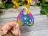 Colorful teardrop-shaped sticker with mystical designs held by a hand on a wooden surface with flowers in the background.