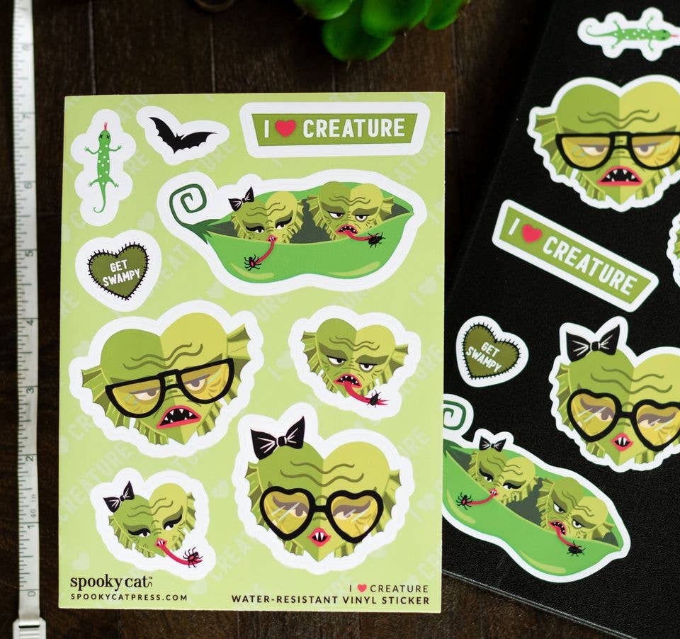 A sheet of cute monster stickers on a light green background, displayed next to a black sheet of stickers on a dark wooden table. The stickers feature a green monster with glasses, bows, and a heart-shaped face, along with text like &
