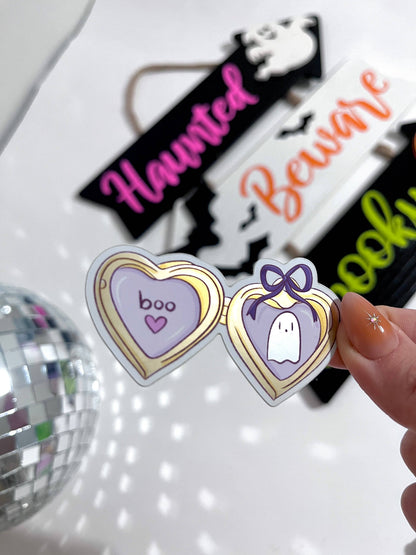 A hand holds a pair of novelty heart-shaped sunglasses with a purple and gold frame, featuring &