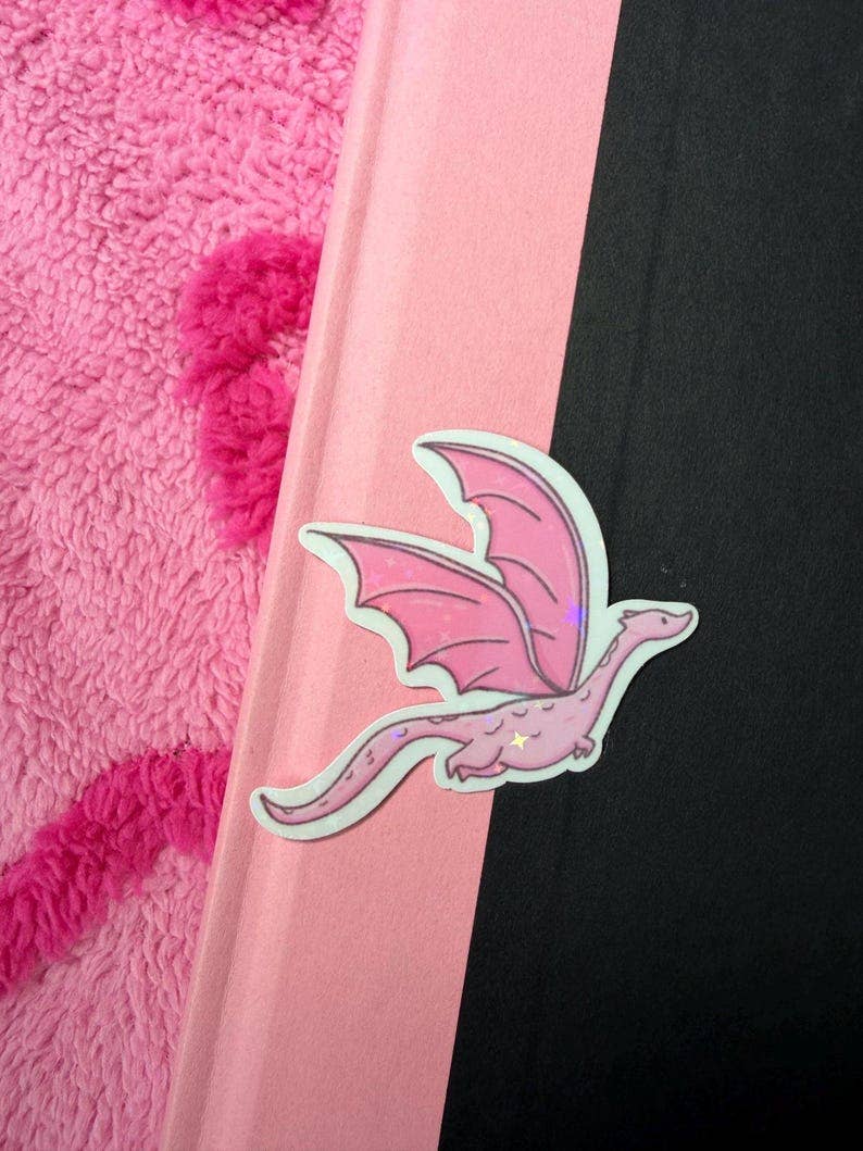 A pink dragon-shaped sticker with white and purple accents, shown resting on a pink and black background next to a fuzzy pink fabric.