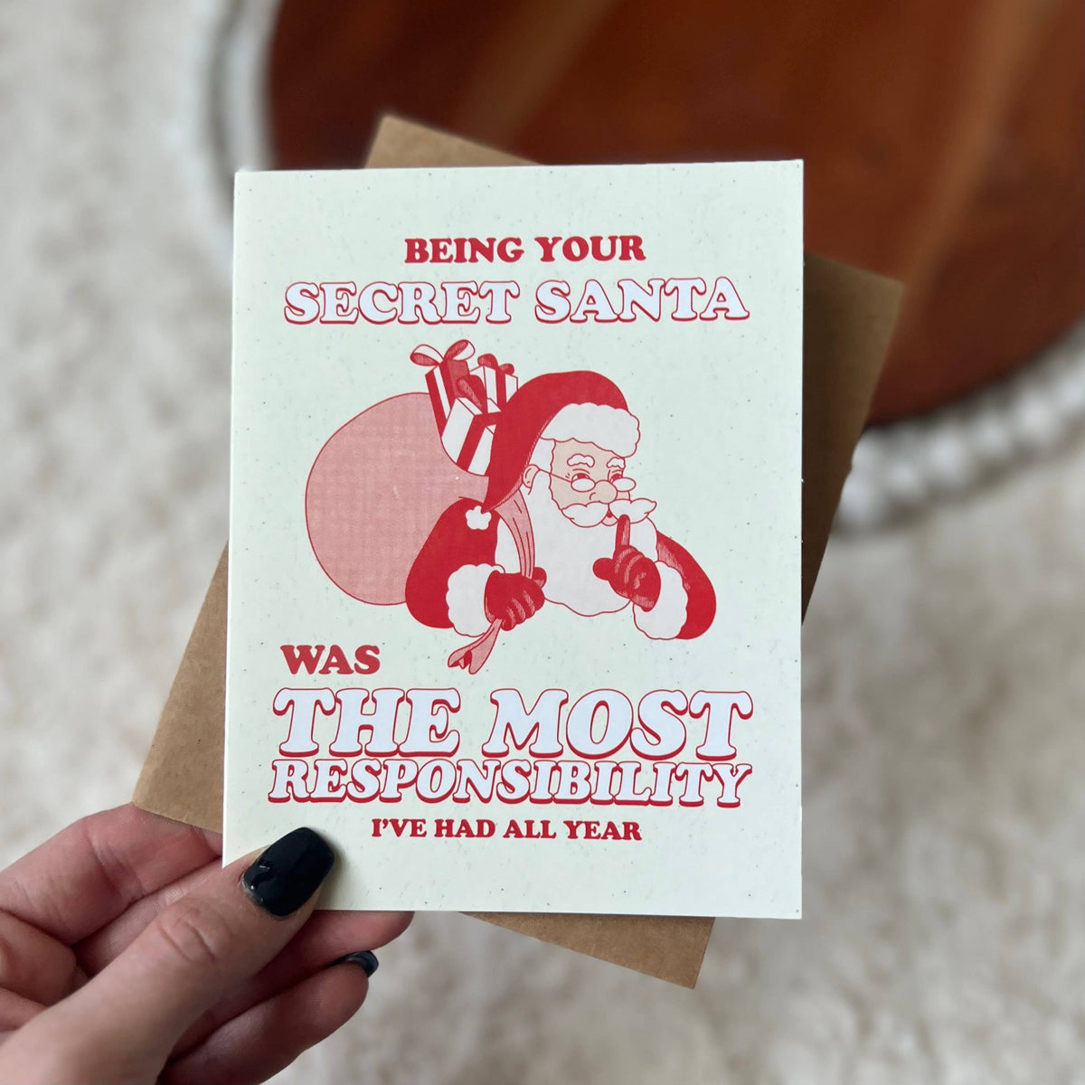 A hand with black nail polish holds a white greeting card featuring a red illustration of Santa carrying gifts, with the text &