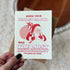 A hand with black nail polish holds a white greeting card featuring a red illustration of Santa carrying gifts, with the text &
