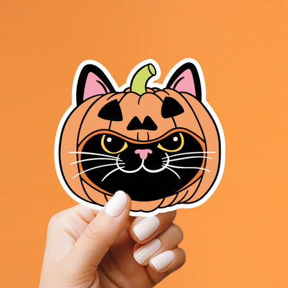A hand with white manicured nails holds a die-cut sticker shaped like a pumpkin-headed cat face. The sticker features an orange pumpkin body with black facial features, pink inner ears, yellow eyes, and white whiskers, set against a solid orange background.