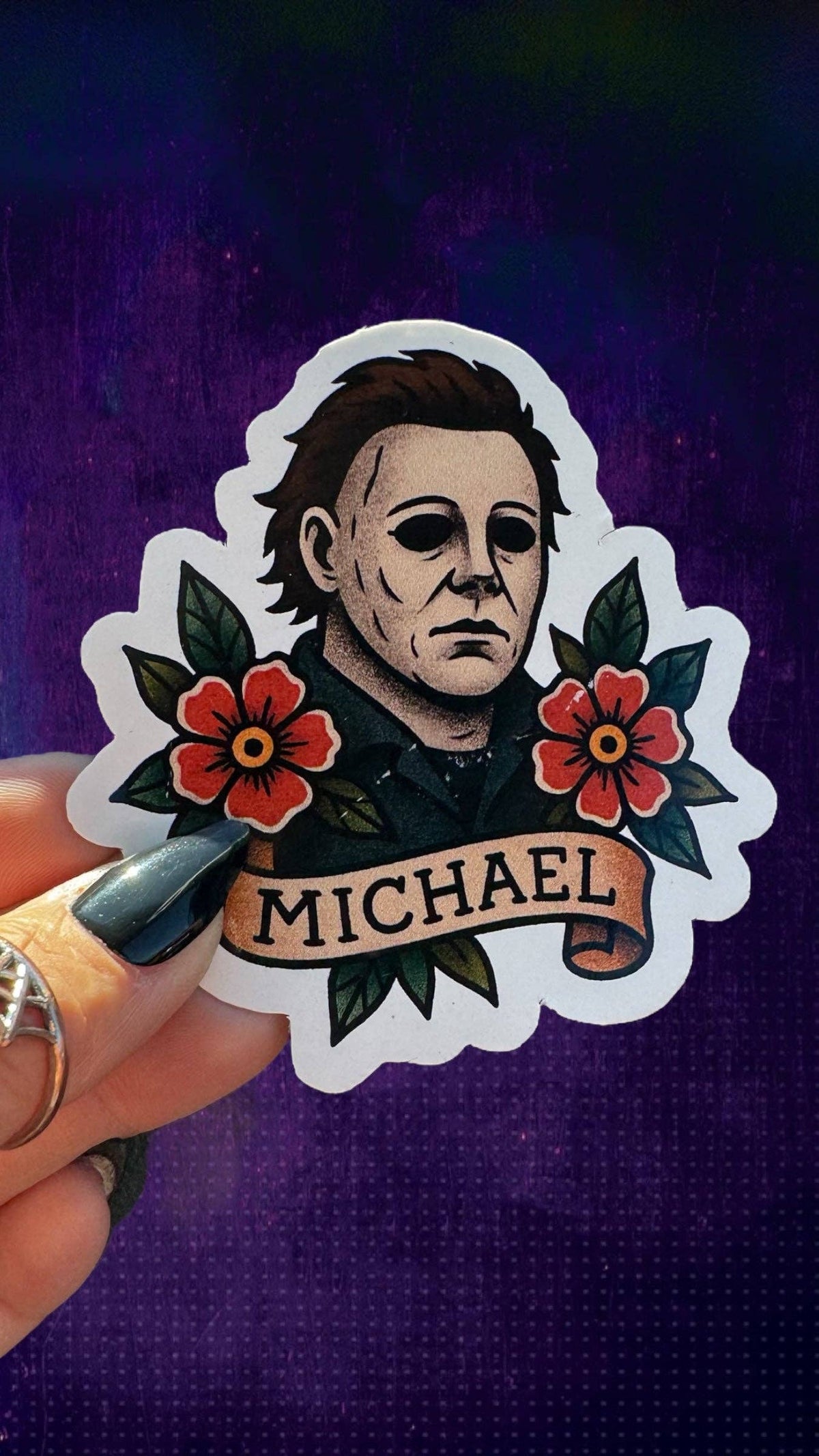 A hand holds a die-cut sticker featuring a portrait of Michael Myers with red flowers and the name &